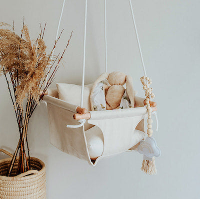 Cuddle bunny sitting in beige baby swing with white ropes and wooden dowels.